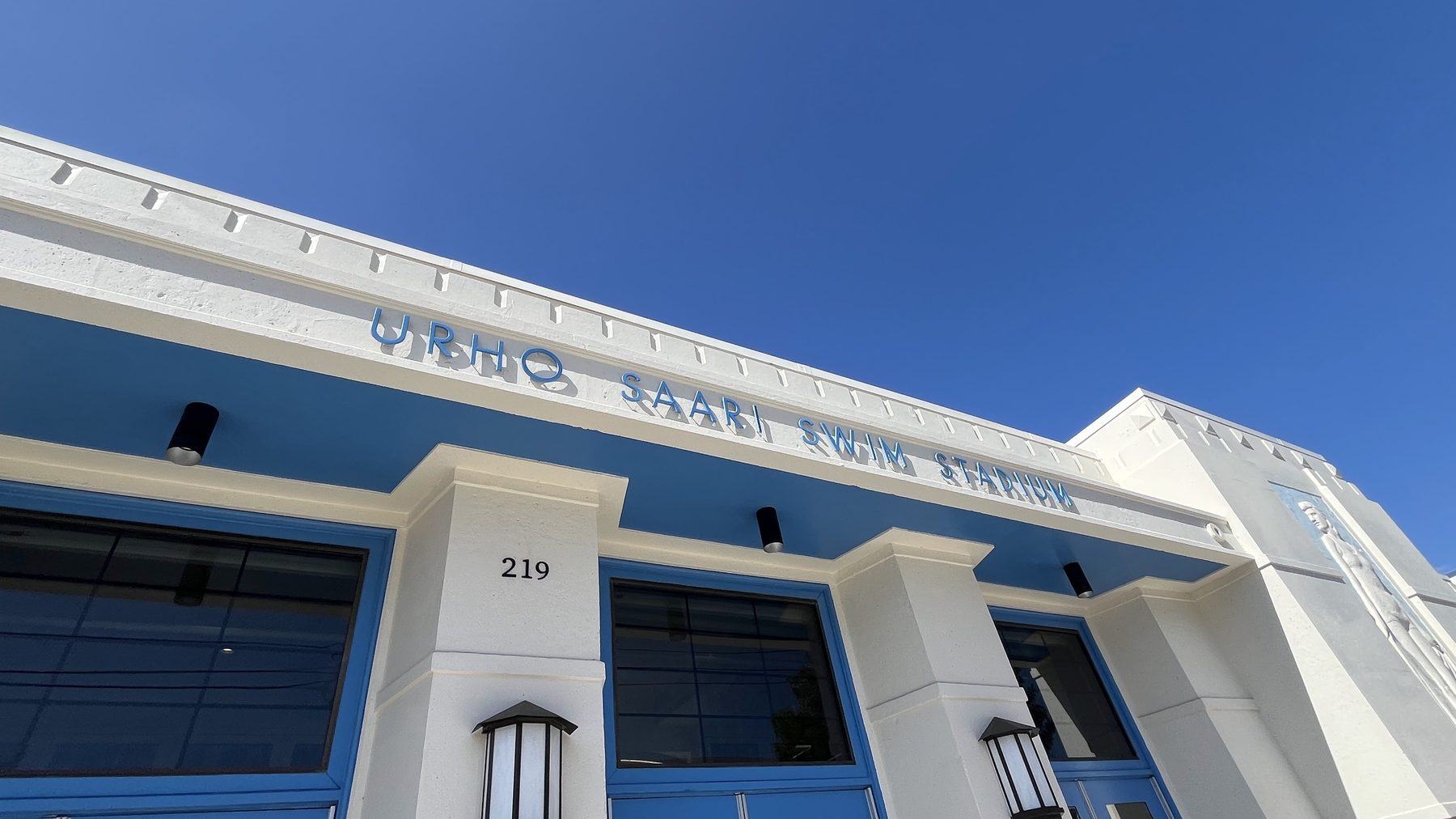 The front of the Urho Saari Swim Stadium, its name in raised letters across the blue and white facade