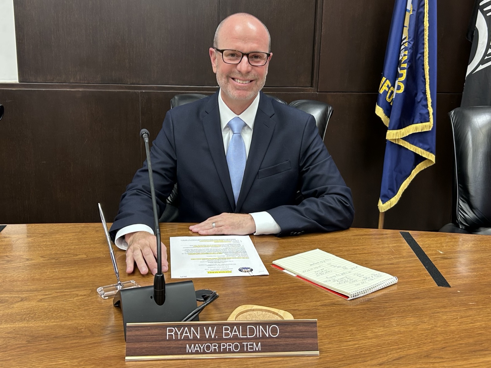 Ryan Baldino seated at the council dais in a dark suit and blue tie, a microphone and papers in front of him and a name plate reading RYAN W. BALDINO, MAYOR PRO TEM