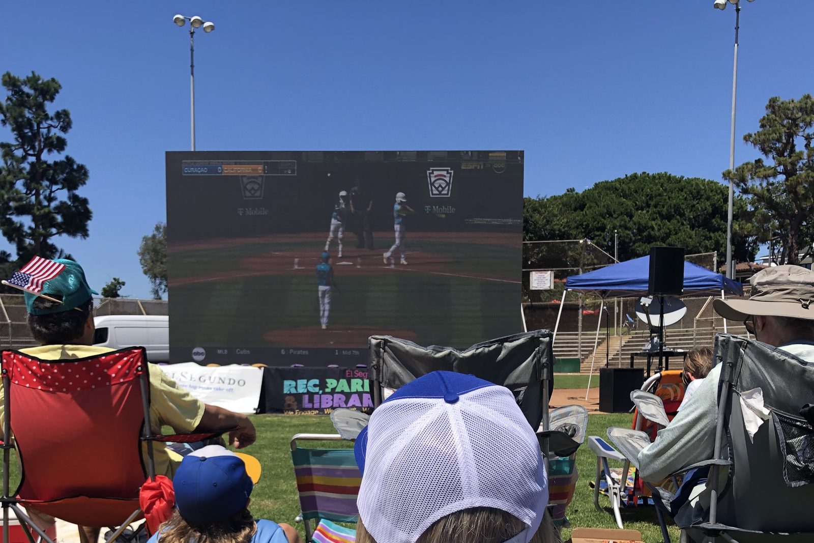 A crowd watching the Little League World Series on the Jumbotron at George Brett Field, seated on blankets and camp chairs.