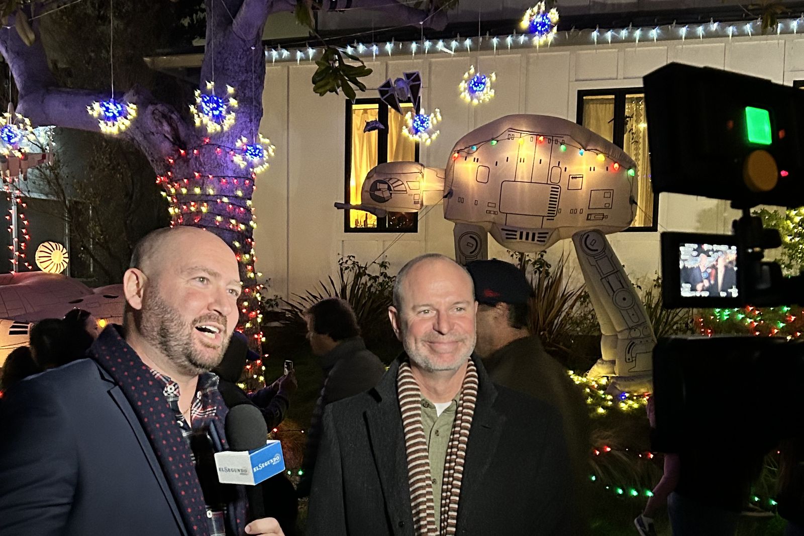 Ryan Baldino being interviewed by a television reporter holding a microphone, in front of a house strung with holiday lights.