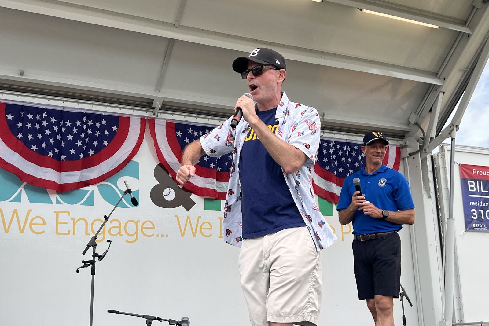Ryan Baldino speaking into a handheld microphone under red, white and blue bunting at a Fourth of July event.