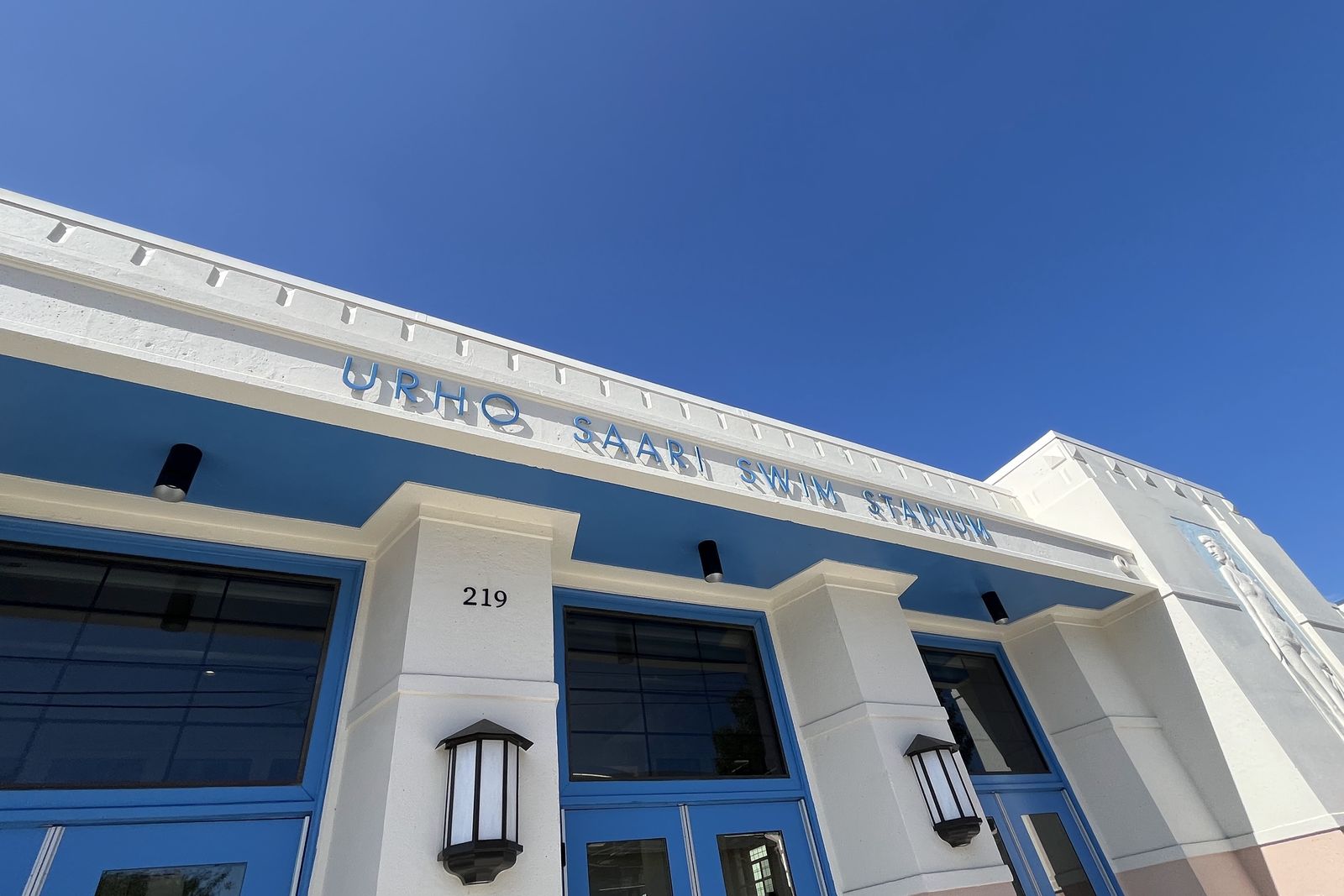 The blue and cream art deco facade of the Urho Saari Swim Stadium with its name in raised lettering.