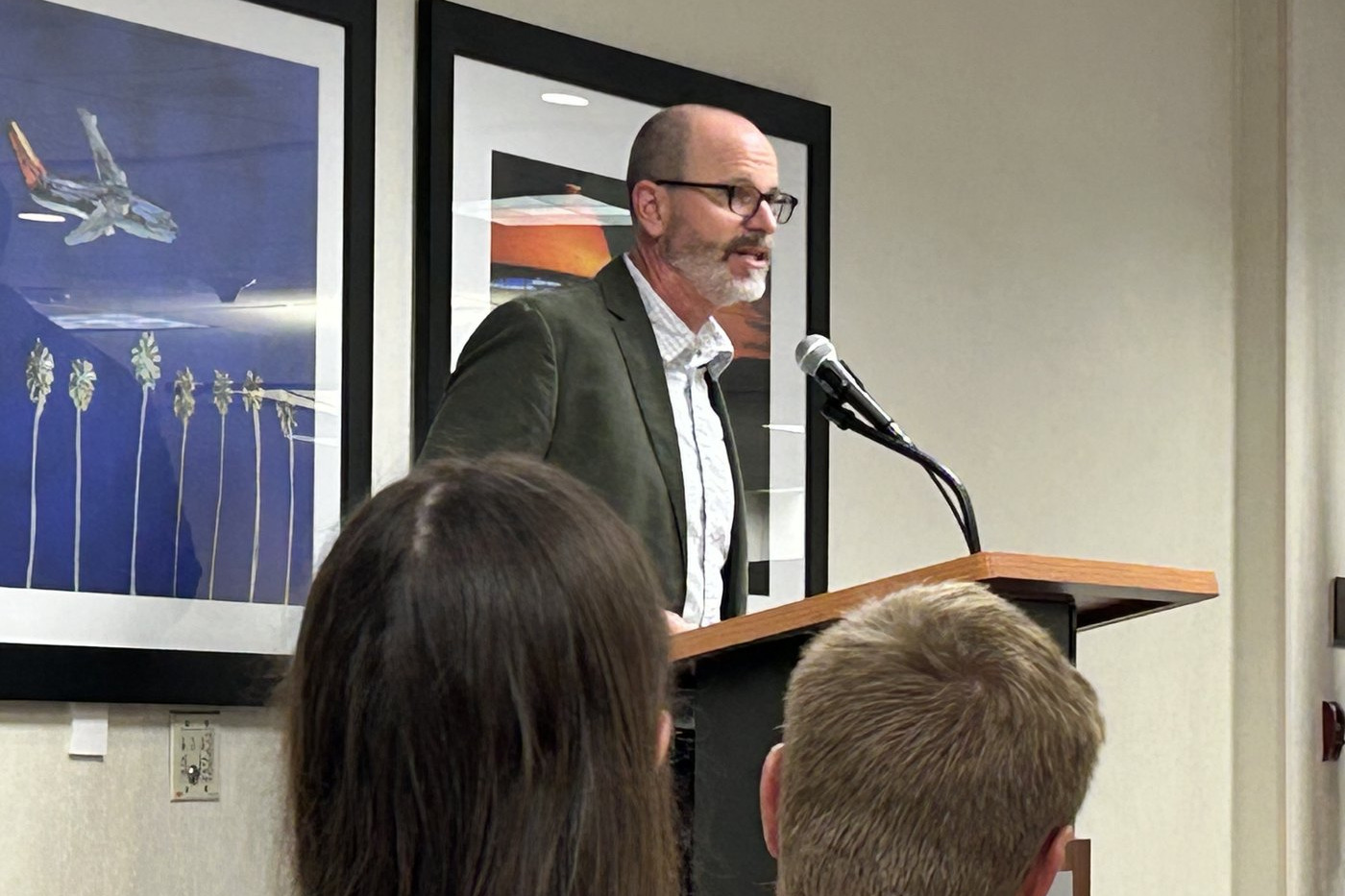 Ryan Baldino speaking at a lectern with a microphone, framed photographs of aircraft on the wall behind him and the backs of two listeners in the foreground